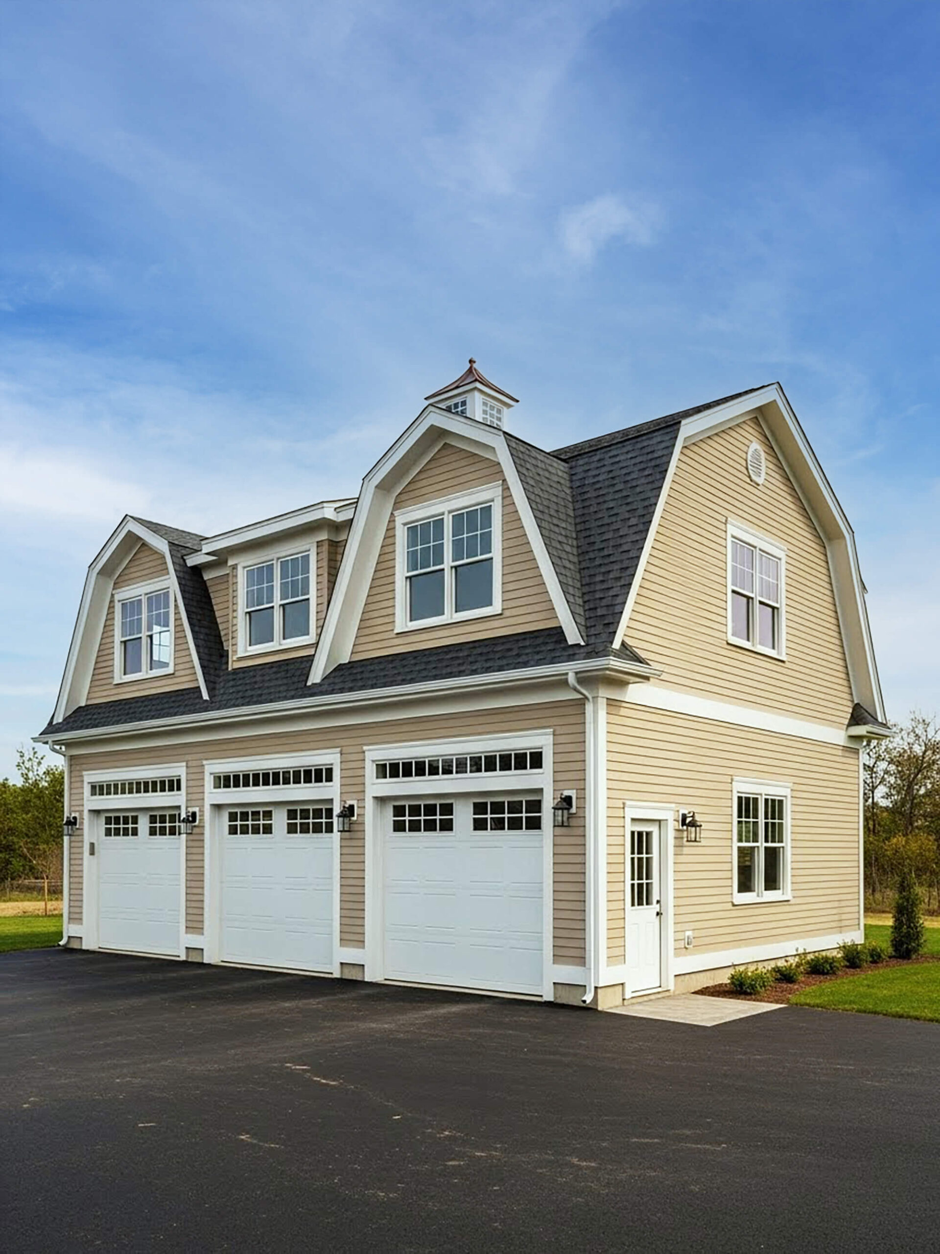 Detached two-story three-car garage with tan siding, white doors and trim, dormer windows, and asphalt driveway.